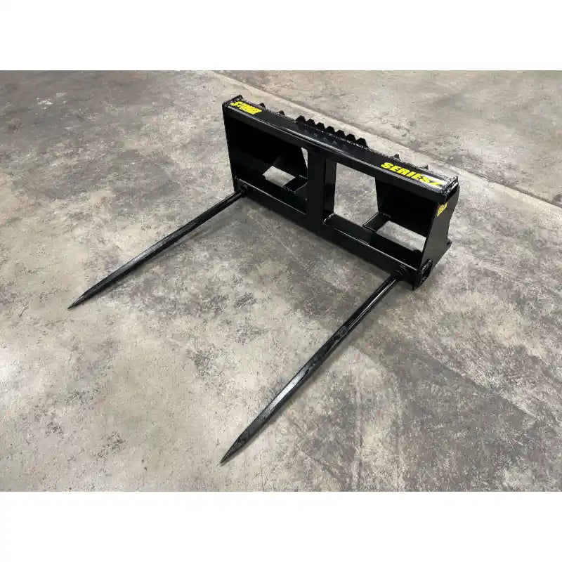 Black steel bale spear attachment with two long pointed tines viewed from front left on concrete floor