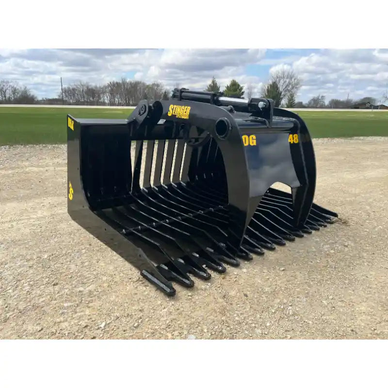 Black steel skid steer rock grapple attachment with spaced curved tines and hydraulic cylinders on gravel surface outdoors