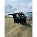 Yellow skid steer loader with open black clam-style grapple attachment holding scrap material over cut grass field