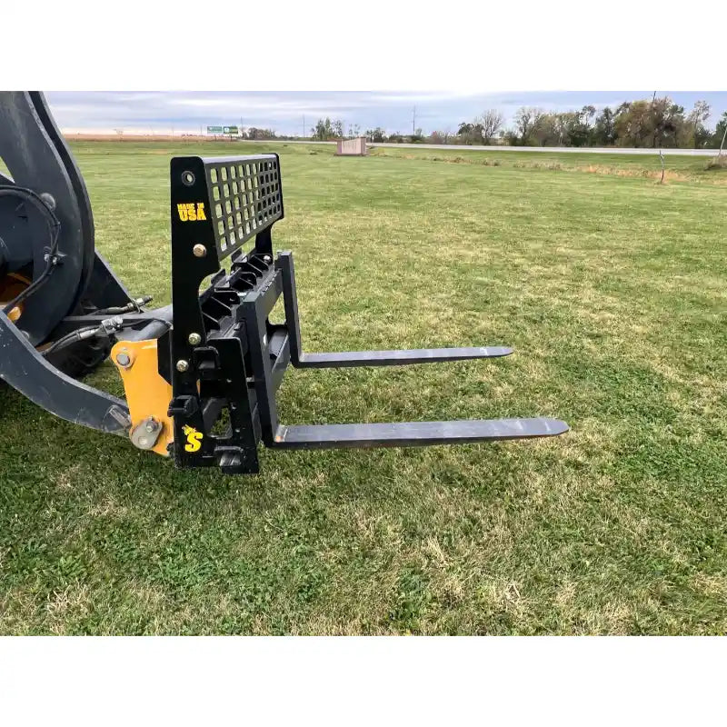 Black skid steer pallet fork attachment with mesh backrest and two straight steel fork tines on grassy field