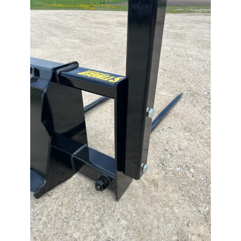 Close-up of black metal skid steer attachment with visible bolt and part of two prongs on gravel surface