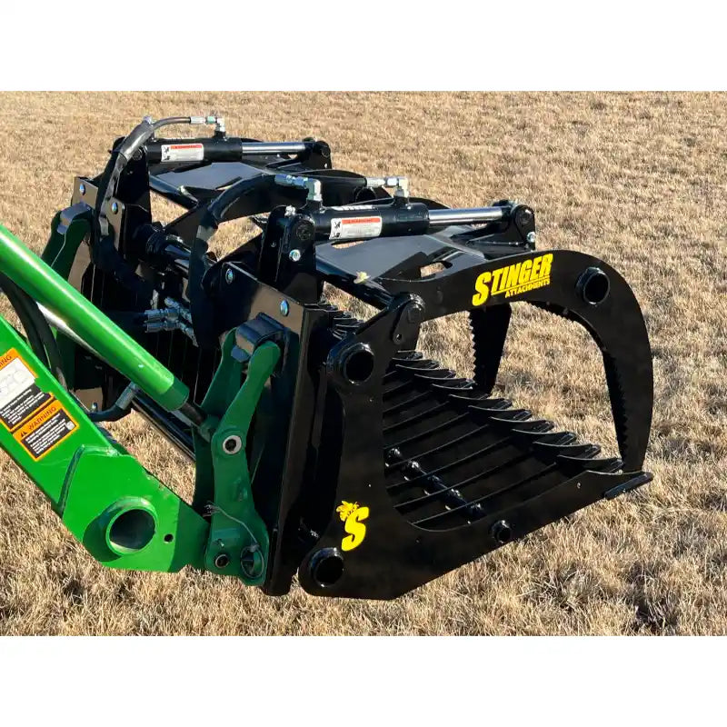 Black steel rock grapple with serrated clamshell jaws and spaced flat tines attached to green tractor loader arm over dry grass