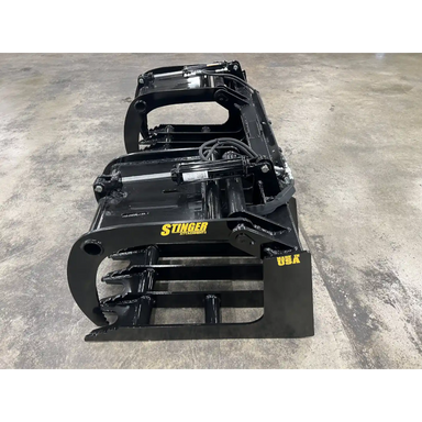 Black skid steer grapple attachment with multiple curved tines, hydraulic hoses, and solid steel frame on concrete floor