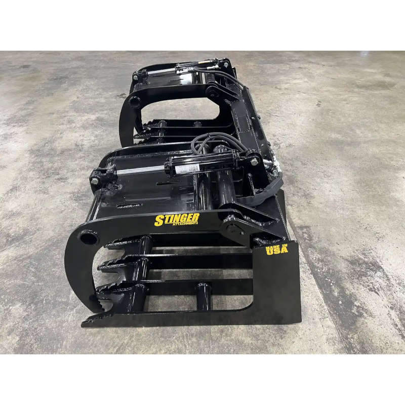 Black painted skid steer grapple attachment with curved tines, hydraulic hoses, and reinforced frame on concrete floor
