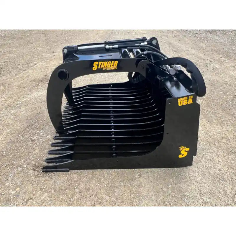Black skid steer rock grapple attachment with spaced curved tines, hydraulic cylinders, and hoses on dirt surface outdoors