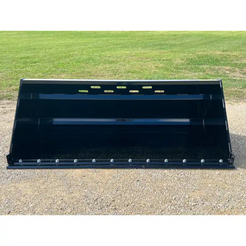 Front view of wide black metal skid steer bucket attachment with bolt-on cutting edge and ventilation slots on gravel surface